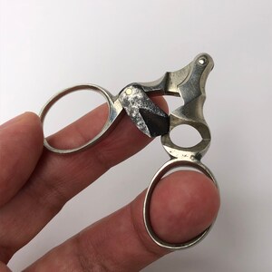 May include: Silver-toned finger ring scissors with a small, round, gold-colored accent. The scissors are open and held by a finger.