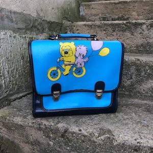 Puede incluir: Una mochila escolar vintage azul con un oso amarillo y una criatura rosa montando una bicicleta con globos. La mochila tiene dos hebillas y una solapa.
