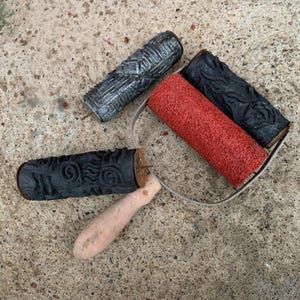 May include: A paint roller tool with a light wood handle and a metal frame holding a red textured roller cover, accompanied by two separate cylindrical patterned rollers, one in black and one in a metallic silver finish.