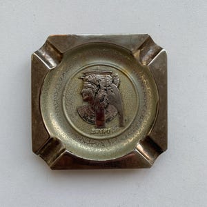 May include: A silver square ashtray with a raised circular design featuring a profile of an Egyptian queen with the word "EGYPT" underneath.
