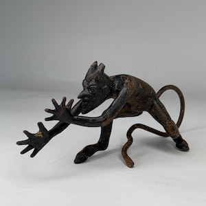 Könnte beinhalten: Eine dunkelbraune Metallskulptur einer Kreatur mit langer Nase, spitzen Ohren und einem eingerollten Schwanz. Die Figur befindet sich in einer hockenden Position mit ausgestreckten Händen, und die Skulptur hat ein strukturiertes, gealtertes Aussehen.