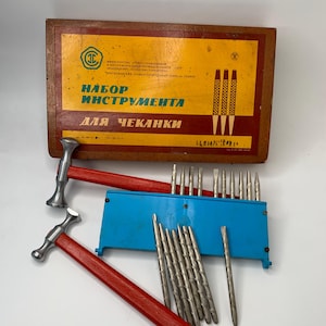 May include: A vintage metalworking set featuring a wooden box with Cyrillic text and a yellow and red design. The set includes two red-handled hammers, a blue tool holder, and various metal chisels and punches. The box reads "Set of Tools for Chasing".