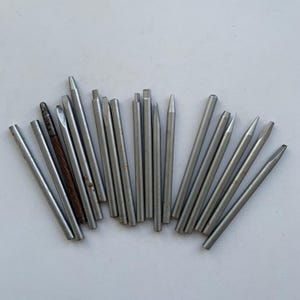 May include: A selection of metal punches and chisels in various sizes and shapes. The tools are silver and arranged on a white surface. Some have flat, pointed, or bevelled ends, while others are cylindrical.