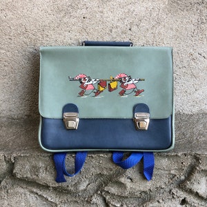 Puede incluir: Una mochila escolar vintage azul y verde con una ilustración de dibujos animados de tres figuras que llevan un palo y bolsas. La mochila tiene dos correas ajustables y un asa superior.