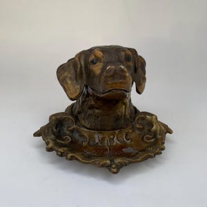 May include: A vintage brass dog head inkwell with a decorative base. The dog's head is brown and the base is gold with a floral design.