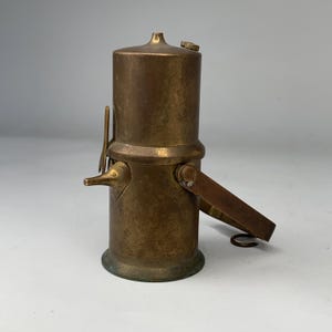 Può includere: Una caffettiera antica in ottone con corpo cilindrico, beccuccio e manico. La caffettiera vintage ha un piccolo coperchio e una leva. L'ottone presenta una calda patina invecchiata. Un classico articolo per la cucina.