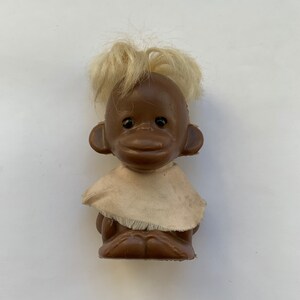 Könnte beinhalten: Eine braune Plastik-Affenpuppe mit blonden Haaren, die ein weißes Stoffkleid trägt. Die Puppe hat schwarze Augen und eine braune Nase.