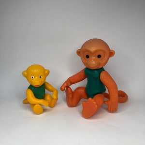 Puede incluir: Dos juguetes de mono de plástico, uno es naranja y el otro es amarillo. Ambos monos llevan camisas verdes y tienen ojos negros.