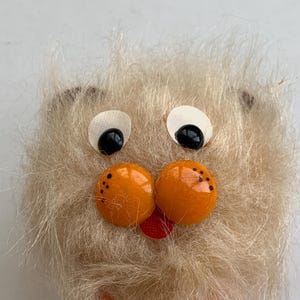 May include: Close-up of a handmade, fuzzy, light brown creature face. It has large white and black eyes, two orange button-like cheeks with black dots, and a small red tongue. The creature's fur is soft and textured.