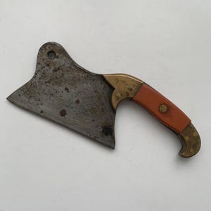 May include: A vintage cleaver with a worn, dark metal blade and a small, orange handle. The handle is accented with brass-colored metal. The cleaver has a hole near the top for hanging.