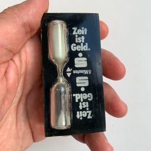 Puede incluir: Un reloj de arena negro con bulbos plateados y el texto "Zeit ist Geld. 8 Minuten. Zeit ist Geld." impreso en la parte delantera.