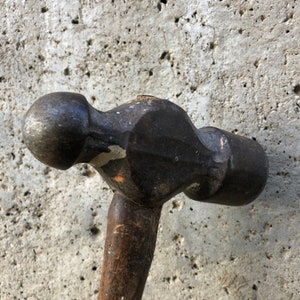 Puede incluir: Un martillo vintage negro y plateado con mango de madera. El martillo tiene una cabeza redonda con una pequeña abolladura en el lateral.
