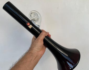 Op de afbeelding: Een zwarte glazen vaas met een uitlopende voet en een lange, cilindrische body. Een helder, gedraaid glazen accent is aan de zijkant bevestigd. Een klein label is zichtbaar op de vaas. De vaas wordt door een persoon vastgehouden.