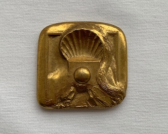 May include: A gold-colored metal plaque with a raised design of a scallop shell, a sphere, and stylized waves. The plaque has a rectangular shape with rounded corners.