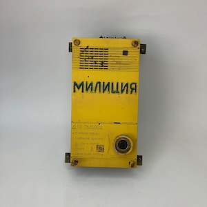 Può includere: Una scatola di metallo gialla con la parola "Milicia" in lettere blu. La scatola ha un pulsante e istruzioni per l'uso in russo: "Per chiamare", "Premere il pulsante", "Indicare il motivo", "Chiamata e numero del vagone", "Rilasciare il pulsante".