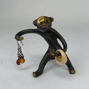 May include: A bronze-colored figurine of a monkey-like figure holding a pair of earrings with amber-colored beads and a cream-colored ring. The monkey has a gold-colored face and feet.