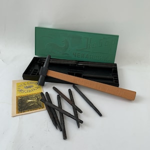 Pode incluir: Um conjunto de trabalho em metal vintage com um martelo com cabo de madeira, vários punções de metal e uma caixa verde com texto cirílico. O conjunto inclui um pequeno livreto com texto cirílico e uma ilustração de veado. O martelo tem uma cabeça preta e um cabo castanho claro.