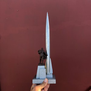 Könnte beinhalten: Eine silberne Modellrakete steht auf einem grauen Sockel mit einer kleinen Bronzestatue einer Person, die auf einer Plattform davor steht.