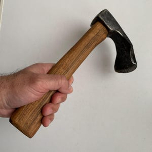 May include: A small, vintage axe with a dark metal head and a wooden handle. The handle is a light brown color with visible wood grain. The axe head has a curved blade and a flat striking surface.