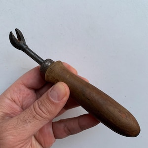 May include: An antique upholstery tool with a dark wooden handle and a metal head. The metal head has a forked design. The tool is held in a hand against a white background. The handle is a dark brown color.