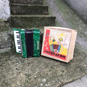 May include: A green accordion with a black bellows and a box with a colourful illustration of children playing accordions. The box reads "Детский Аккордеон".
