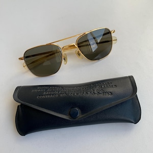 May include: Gold-framed aviator sunglasses with dark lenses rest above a black leather case. The case has a snap closure and embossed text. The sunglasses have a double bridge and a classic design.