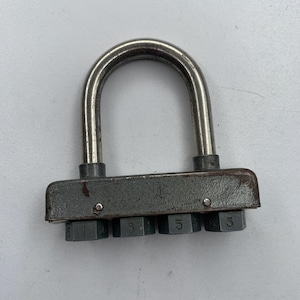 Könnte beinhalten: Ein graues Zahlenschloss mit einem silbernen Metallscharnier und sechs nummerierten Zifferblättern. Die Zahlen auf den Zifferblättern sind 6, 1, 3, 3, 5 und 5.