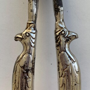 Può includere: Primo piano di uno schiaccianoci d'argento vintage. Le maniglie sono a forma di aquile con dettagliate decorazioni di piume e becco. Il metallo mostra segni di età e usura, aggiungendo al suo fascino antico. Un classico utensile da cucina.