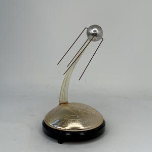 Puede incluir: Un modelo de Sputnik plateado con una parte superior esférica y cuatro antenas. El modelo tiene un soporte transparente y curvado y una base con texto grabado y un borde negro. El texto en la base dice "4X-1957 CCCP".