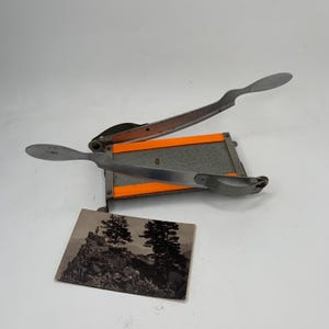 May include: A grey paper cutter with a silver blade and orange accents. A black and white photograph of a mountain scene with two trees is next to the cutter.