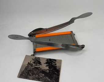 May include: A gray paper cutter with a silver blade and orange accents. A black and white photograph of a mountain scene with two trees is next to the cutter.