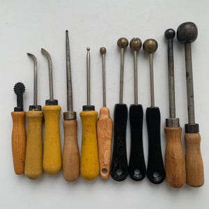 May include: A collection of vintage metal tools with wooden and black handles. The tools include various shapes, such as curved blades, pointed tips, and ball-shaped ends. The handles are in shades of yellow, brown, and black.