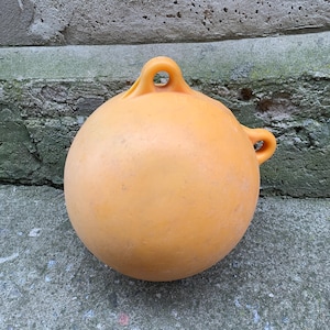 Pode incluir: Uma bóia laranja brilhante com uma alça de laço. A bóia é feita de plástico e tem uma superfície lisa e arredondada.
