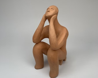 May include: A terracotta sculpture of a person sitting with their chin resting on their hands, looking upwards.