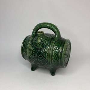 May include: A green ceramic decanter shaped like a barrel with intricate floral designs. The decanter has a handle and a spout, and it sits on three small feet.