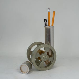 May include: A silver-colored desk organizer set. The set includes a cylindrical pencil holder with pencils and pens, two decorative wheels, and a small cylindrical container. The wheels have a unique, open design.