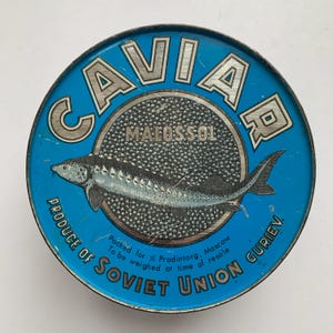 May include: A round, blue tin of caviar with the word "CAVIAR" in silver lettering. The tin features a detailed illustration of a sturgeon and the text "MALOSSOL" and "PRODUCE OF SOVIET UNION GURIEV".