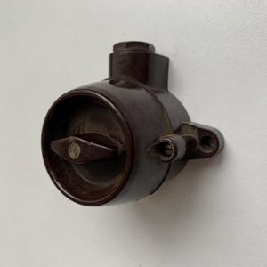 May include: A vintage brown electrical switch with a circular body and a diamond-shaped lever. The switch has a small, rectangular mounting bracket with two holes. The lever has a small, circular emblem.