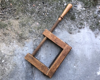May include: A vintage wooden C-clamp with a threaded rod and a wooden handle. The clamp is a warm brown color and appears to be well-used. The background is a mottled gray concrete surface.