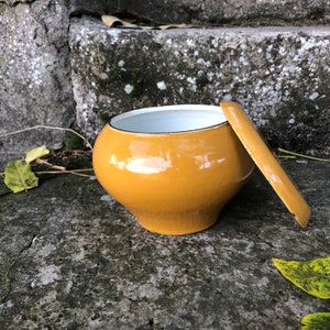 Könnte beinhalten: Ein gelber Emailletopf mit weißem Innenraum und passendem Deckel. Der Topf steht auf einer Steinoberfläche.