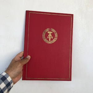 May include: A red, hardback book with a gold emblem on the front cover. The emblem features a compass, hammer, and wheat stalks.