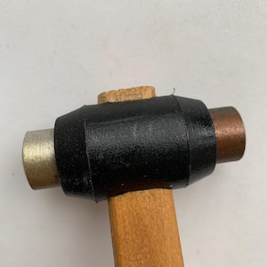 May include: A small, wooden mallet with a black rubber head and a light brown wooden handle. The mallet has a metal striking surface on one end and a copper striking surface on the other.