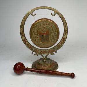 May include: An antique brass gong with a decorative floral design and a wooden mallet. The gong has a circular frame and a round, decorated face. The mallet is dark brown with a rounded head. The gong is approximately 10 inches in diameter.
