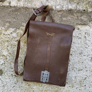 May include: A brown leather messenger bag with a shoulder strap and a metal clasp. The bag features a flap closure and a small gold emblem of crossed swords. The bag is rectangular and appears to be vintage.