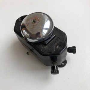 Puede incluir: Un timbre eléctrico negro con una campana cromada y una pequeña perilla en el lateral. El timbre tiene el texto "121121" impreso en él.