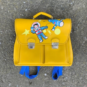 Puede incluir: Una mochila escolar vintage amarilla con un diseño de astronauta azul y rojo. La mochila tiene dos compartimentos grandes y dos bolsillos más pequeños en la parte delantera. La mochila tiene dos correas azules.