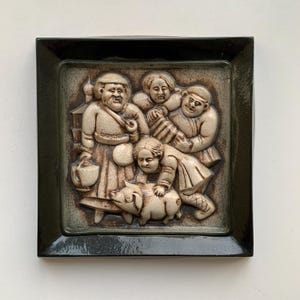 May include: A ceramic wall plaque with a raised relief design of four people and a pig. The plaque is framed in a dark green border. The people are dressed in traditional clothing and are smiling. The pig is standing on its hind legs and is looking up at the people.