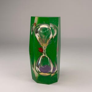 May include: A faceted, translucent green resin block containing an hourglass with purple sand. The resin is embedded with small silver glitter particles and a single red flower, creating a layered, geometric appearance around the central glass timer.