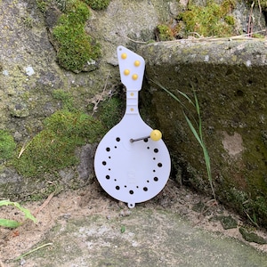 Può includere: Un giocattolo musicale in plastica bianca a forma di chitarra con punti gialli e una piccola manopola. Il giocattolo è appoggiato su una superficie rocciosa ricoperta di muschio.