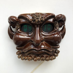 May include: A brown ceramic wall clock shaped like a face with a digital display showing the time as 04:10. The clock has a detailed design with a sculpted beard and horns.